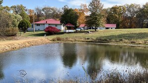 A pond surrounded by a grassy area with a house and a shed in the background. - Peaceful, spacious 4 bedroom house in the country - Gazebo/Pond
 (Keosauqua)