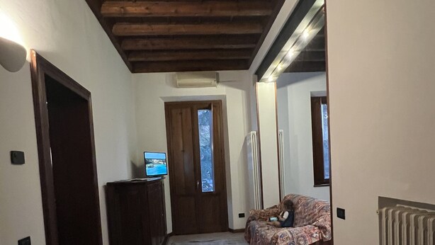 A narrow hallway with a patterned rug, wooden ceiling beams, a door leading to a room with a bed, and a radiator on the wall.