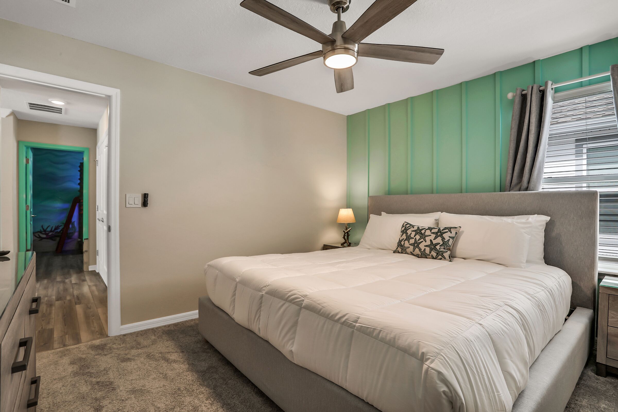Un dormitorio con una cama grande, un ventilador de techo, una mesita de noche con una lámpara y vistas a una habitación con una pared verde y una ventana con persianas.