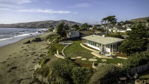 A coastal property with a large house featuring a covered patio, a beach view, and a hilly landscape in the background. - Sunrise to Sunset Views • Cayucos Beachfront Home (Cayucos)