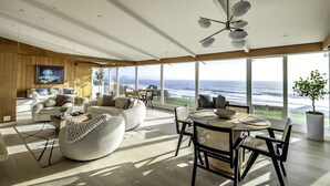 A spacious living room with a large window overlooking the ocean, wooden paneling, and modern furniture. - Sunrise to Sunset Views • Cayucos Beachfront Home (Cayucos)