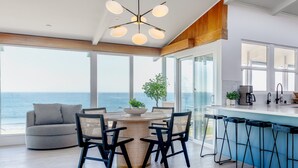 A modern kitchen with a large island, a dining area with a round table and chairs, and a view of the ocean. - Sunrise to Sunset Views • Cayucos Beachfront Home (Cayucos)