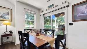 A dining area with a wooden table and chairs, a lamp, and a view of the outdoors through large windows.