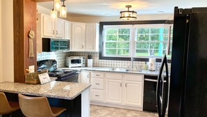 Une cuisine avec des meubles blancs, un plan de travail en granit, un réfrigérateur noir et une fenêtre donnant sur un paysage verdoyant.