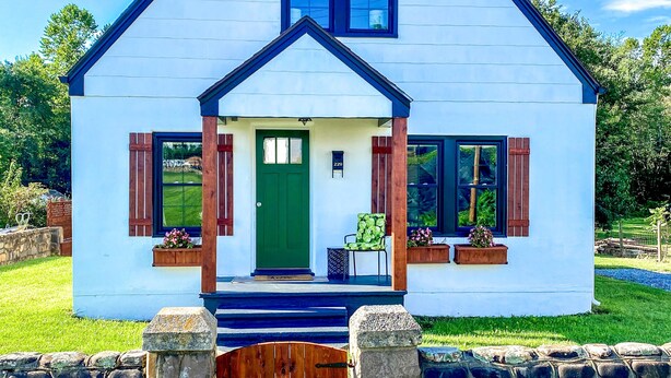 Une maison blanche avec une porte verte, deux fenêtres et un mur de pierre à l’avant.