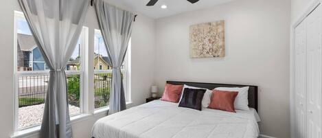 Une chambre à coucher avec un lit, un ventilateur de plafond, une fenêtre avec des rideaux, une table de chevet et un tableau au mur.