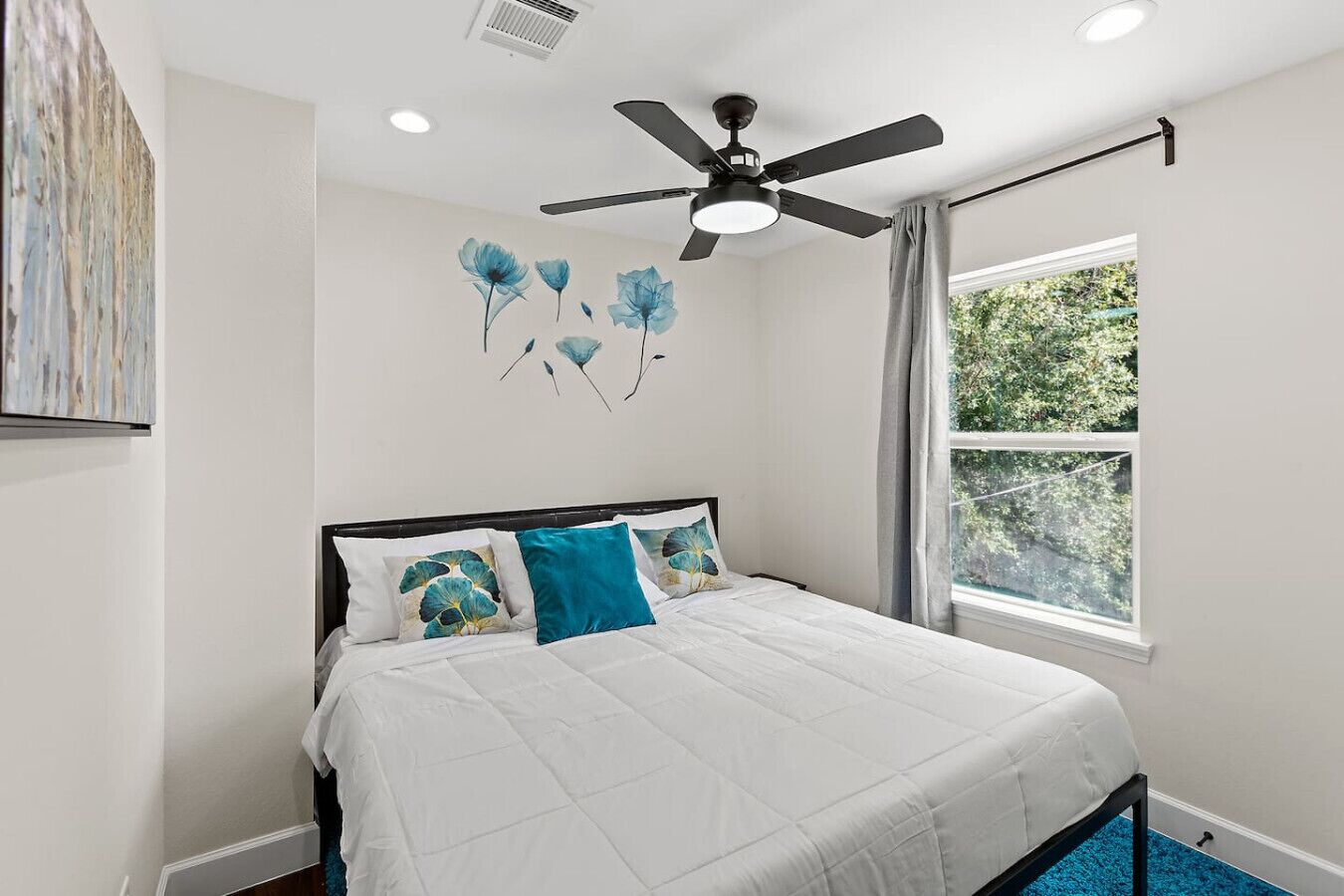 Une chambre à coucher avec un lit, un ventilateur de plafond, une fenêtre avec des rideaux et un mur orné d’une fresque florale.