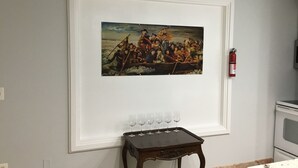 Kehyksiin asetettu historiallinen maalaus seinällä, pieni puinen pöytä, jossa on laseja, ja palosammutin.