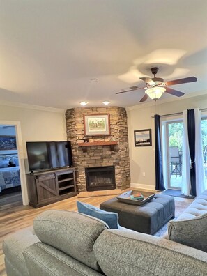 A living room with a stone fireplace, a ceiling fan, and a television.