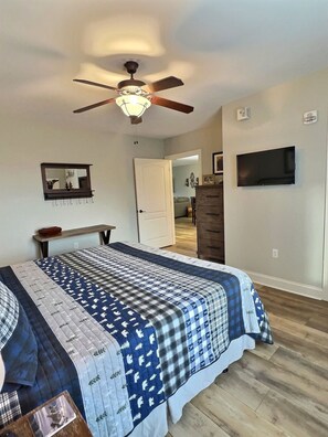 A bedroom with a bed, a ceiling fan, a TV, and a door leading to another room.