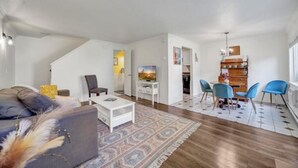 Een moderne woonkamer met een bank, salontafel en een televisie. Er is een eethoek met blauwe stoelen en een kamerkleed met een patroon.