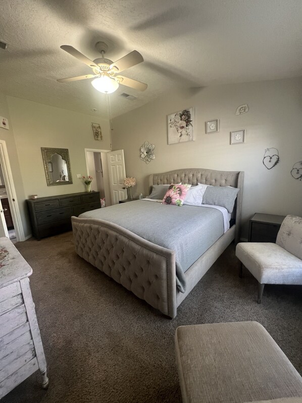 Relax in a serene bedroom with a large tufted headboard bed, ceiling fan, dresser, and vanity with mirror.