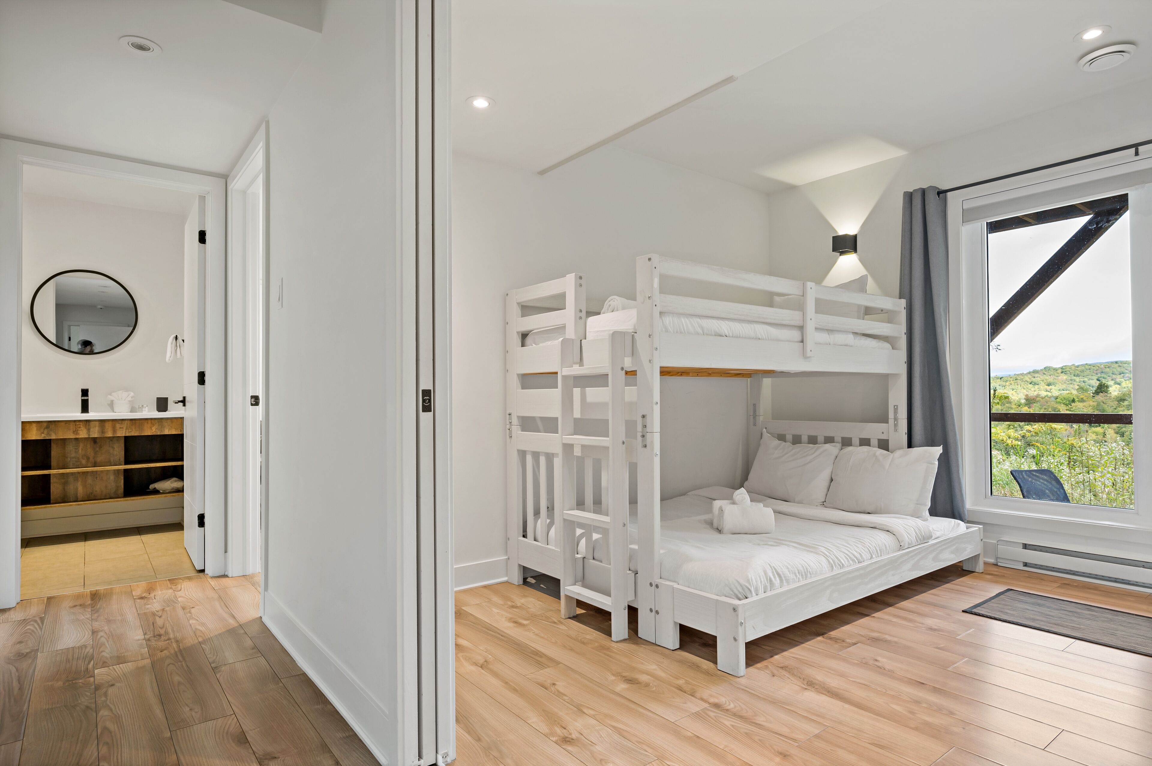 A bedroom with a bunk bed, wooden flooring, a large window with a view, and a bathroom with a mirror and shelves.