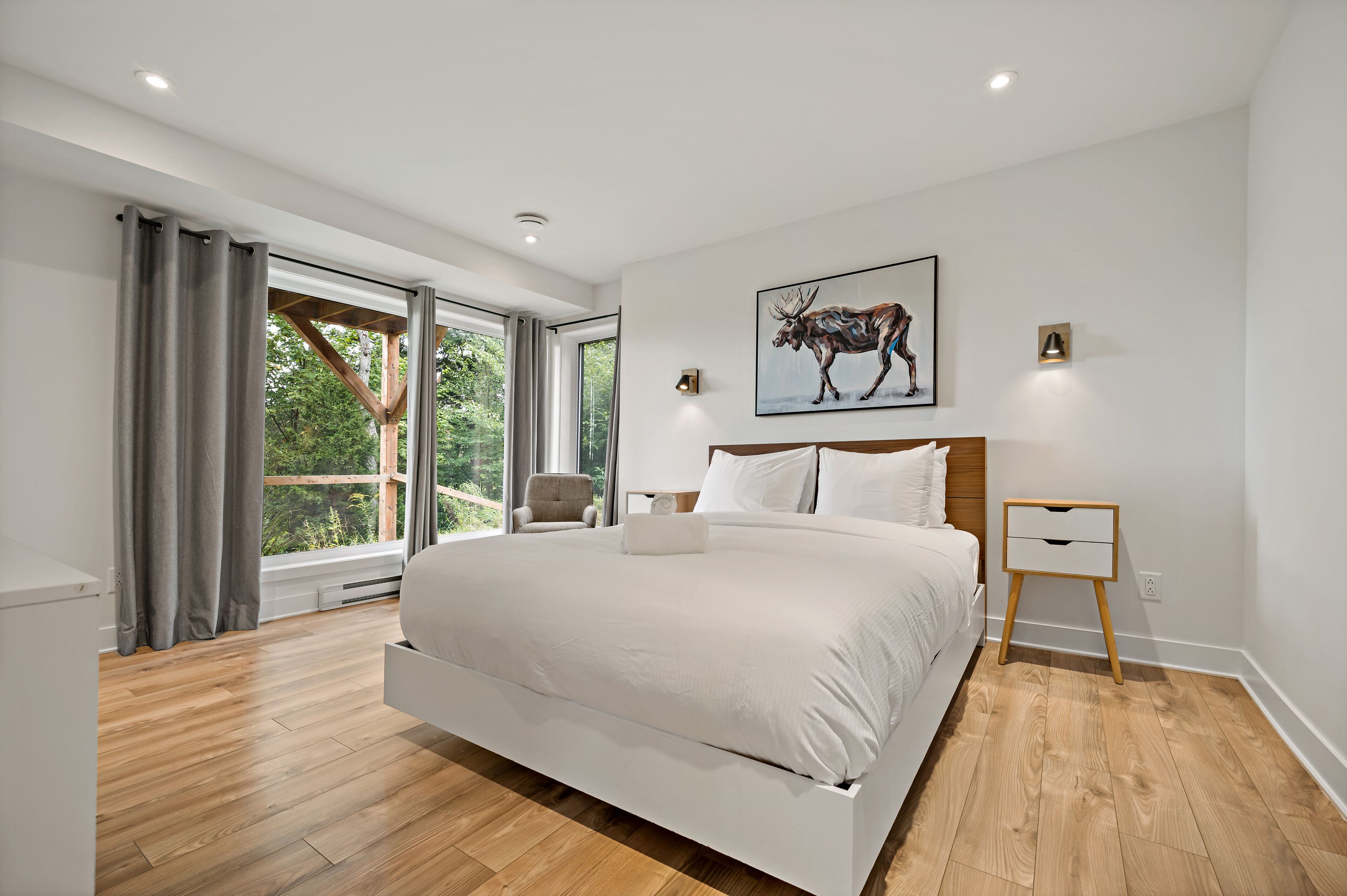 A modern bedroom with a large bed, a wooden floor, a window with curtains, a chair, and a painting of a moose.