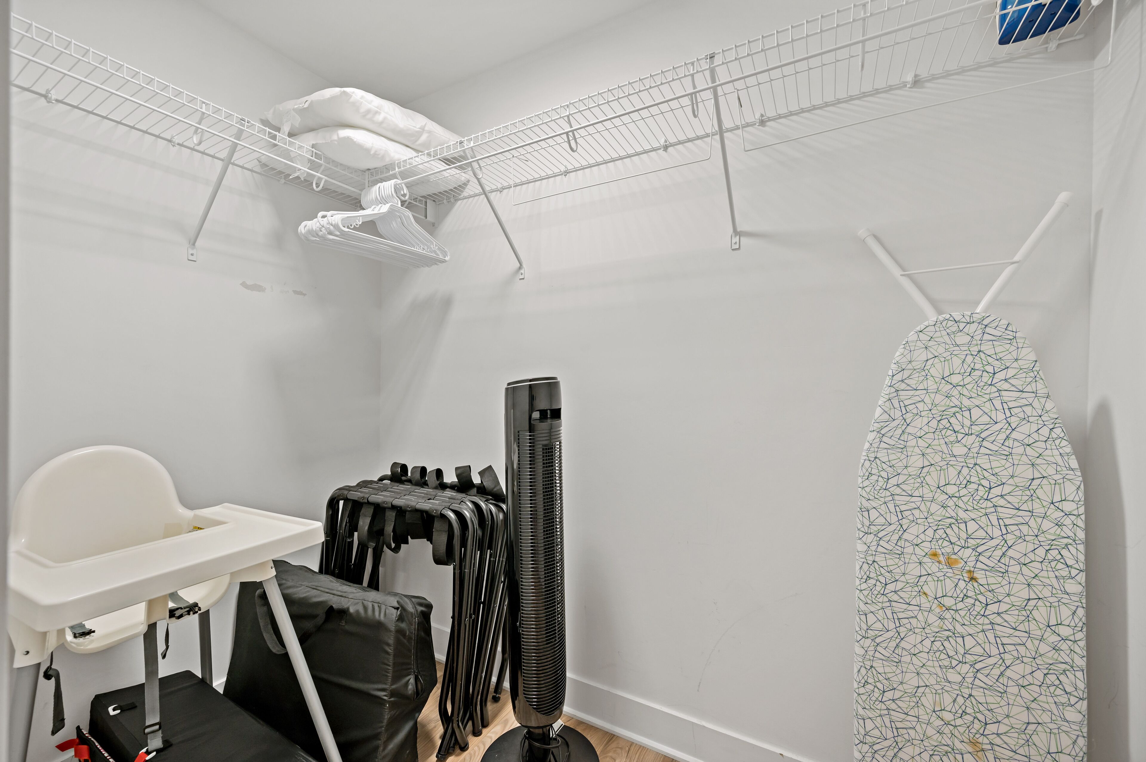 Ein kleines Zimmer mit weißer Wand, einem weißen Stuhl, einem schwarzen Standheizkörper, einem weißen Bügelbrett und einem weißen Regal mit Kleidung.