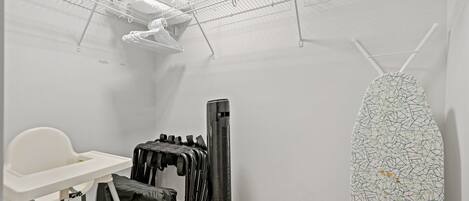 Ein kleines Zimmer mit weißer Wand, einem weißen Stuhl, einem schwarzen Standheizkörper, einem weißen Bügelbrett und einem weißen Regal mit Kleidung.