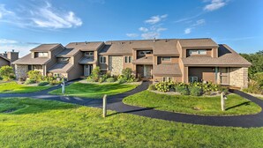 Une rangée de maisons modernes à deux étages, aux façades en pierre et en bois, reliées par une allée commune, et entourées de pelouses et d’arbustes bien entretenus.