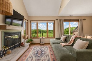 A cozy living room with a fireplace, large windows, a green sofa, and a patterned rug. - Boutique townhome w/ fire pit, KING bed, mezzanine loft & golf views! (Galena)