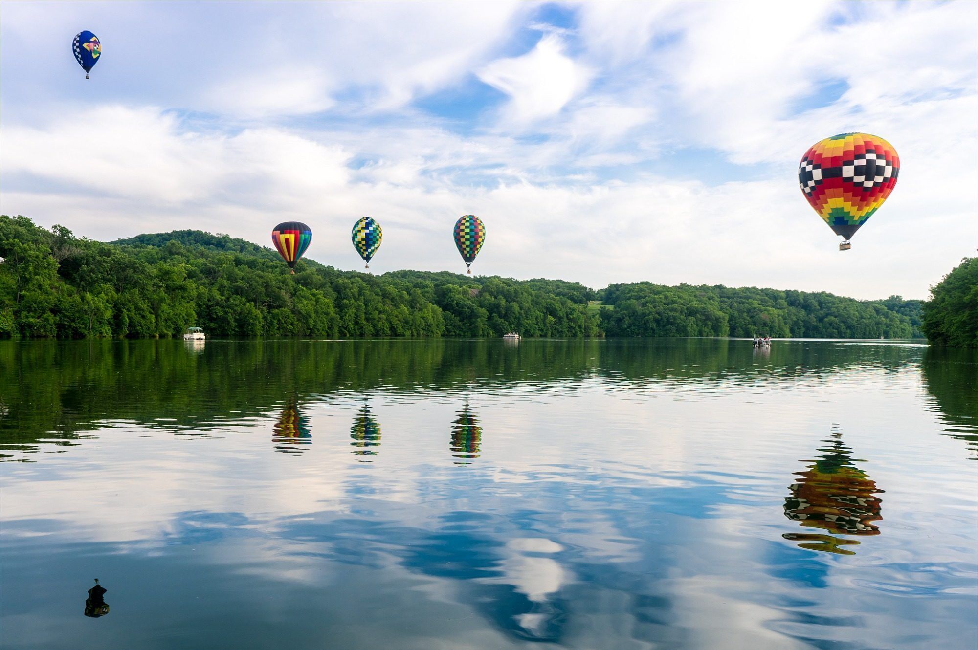 A calm lake with hot air balloons in the distance and a boat on the water.