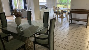 Een eethoek met een glazen tafel en stoelen, een vaas met bloemen, en uitzicht op de omgeving door grote ramen.
