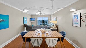 A dining area with a wooden table and white chairs, a ceiling fan, and a view of the ocean through a large window.
