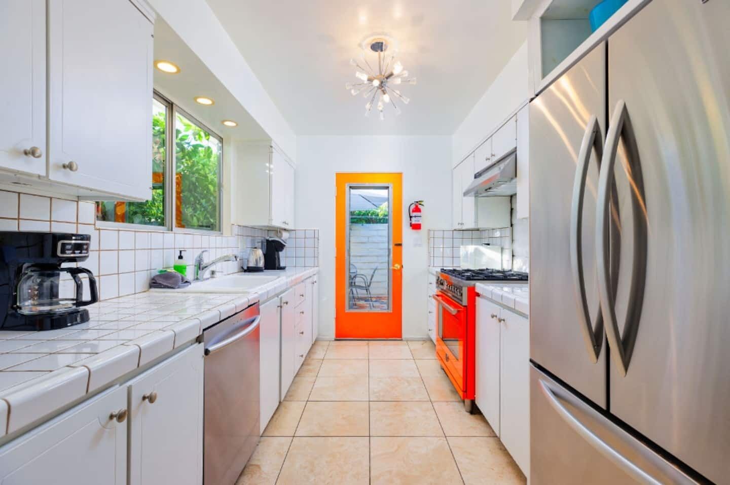 A modern kitchen with white cabinets, a red door, stainless steel appliances, and a tiled floor.