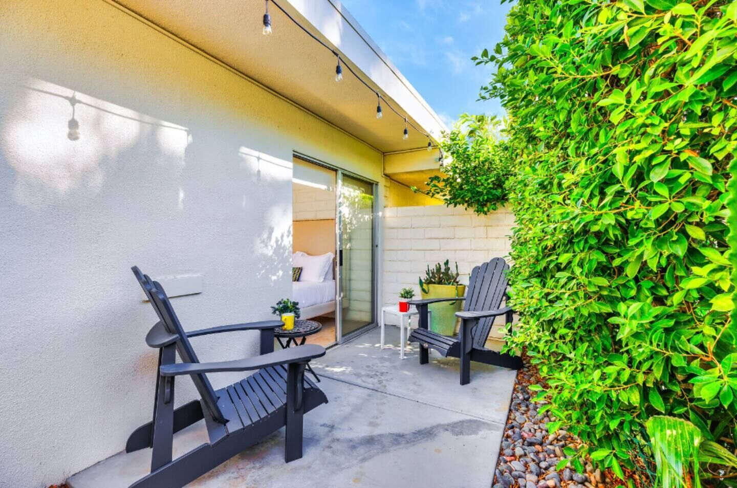 A patio with two chairs and a small table, a potted plant, and a green hedge.