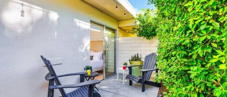 A patio with two chairs and a small table, a potted plant, and a green hedge.