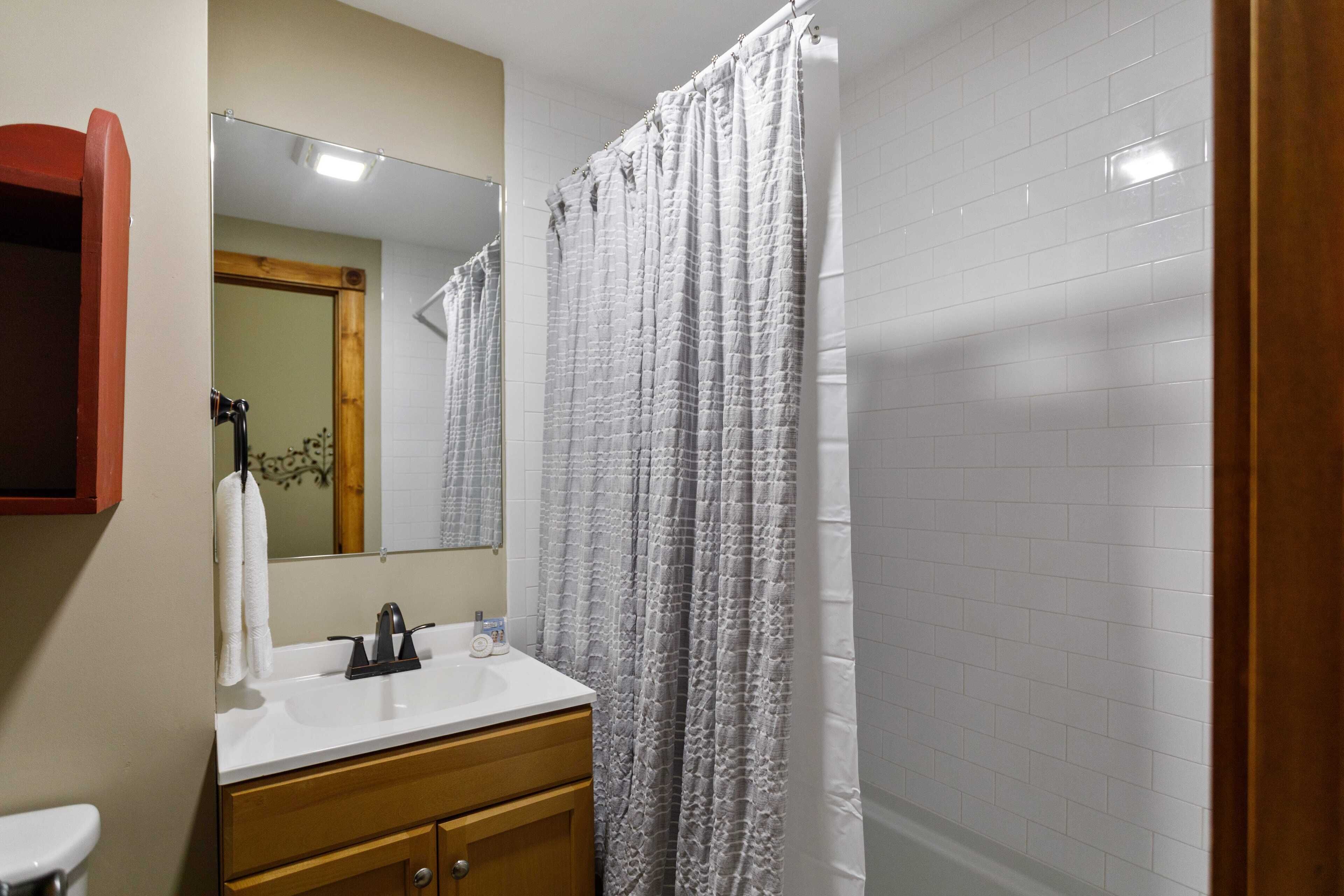 Un baño con pared de azulejos blancos, tocador de madera con lavabo y espejo, y cortina de ducha.