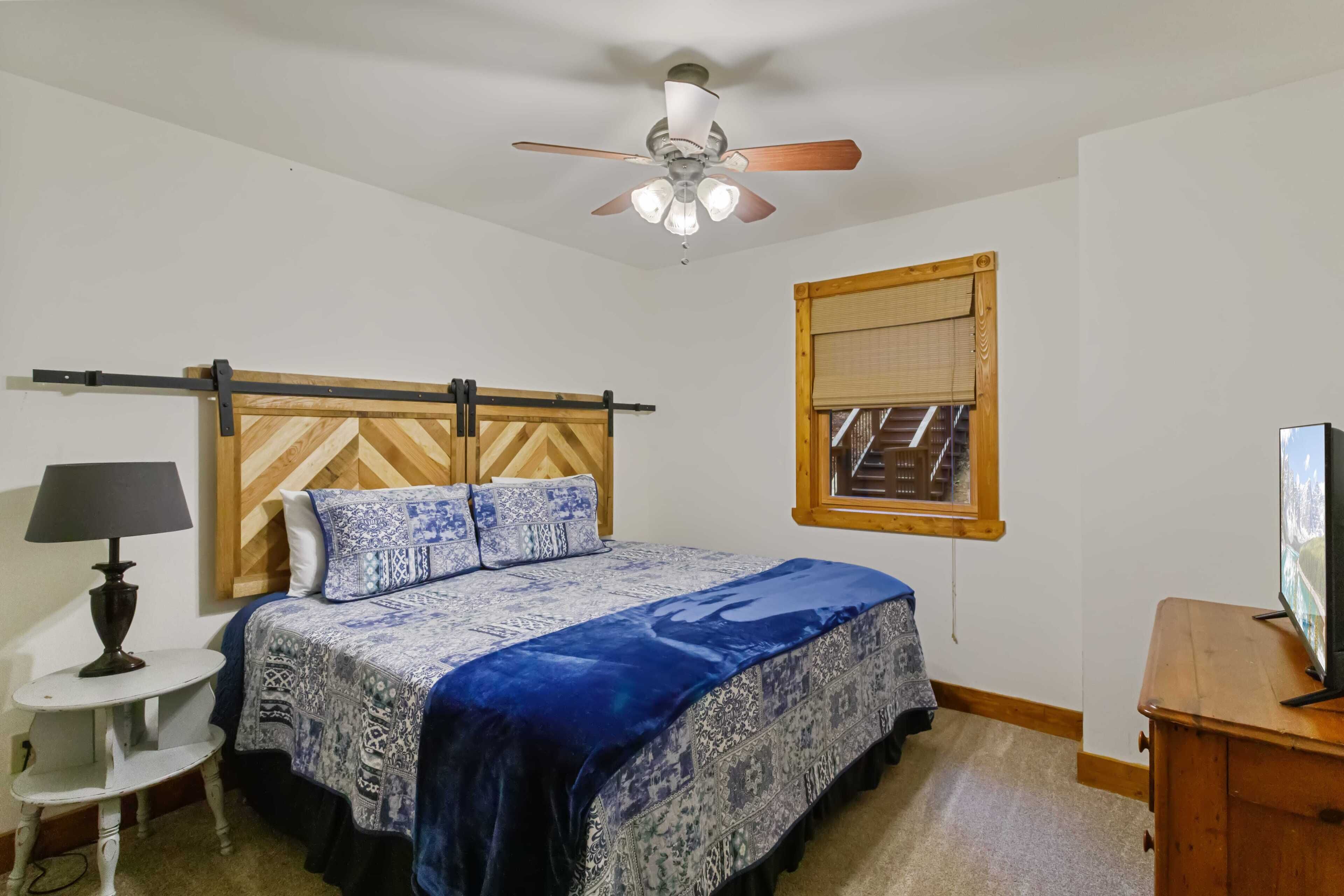 Ein Schlafzimmer mit Bett, Deckenventilator, Fenster mit Jalousien, einem kleinen Tisch mit Lampe und einer Holzkommode.
