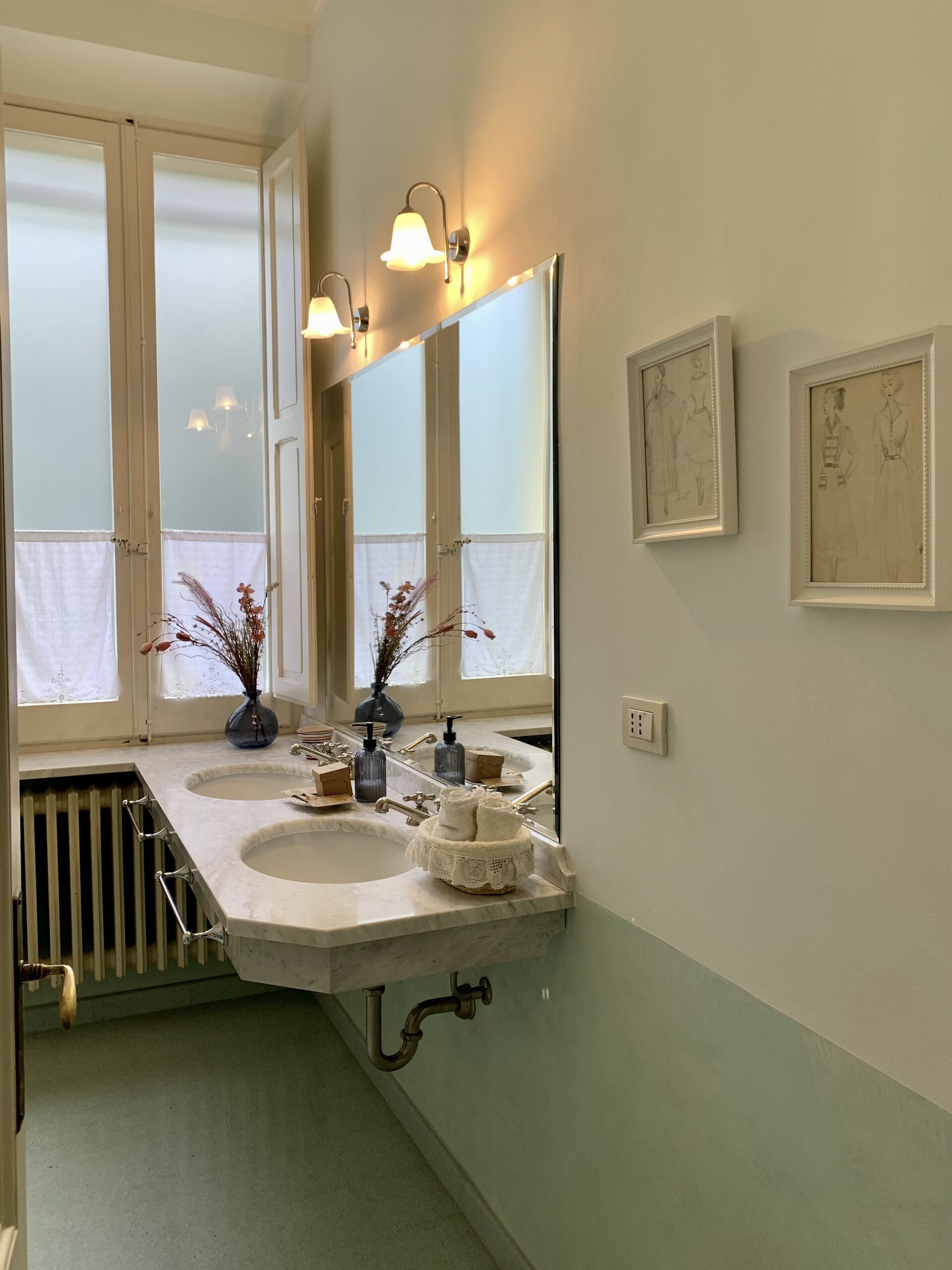 A bathroom with two sinks, a large mirror, and wall-mounted lights. There are framed sketches on the wall.