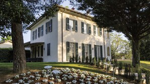 A two-story building with a symmetrical facade, a covered porch, and a well-maintained garden with white flowers. - Villa Monacelli (Fano)