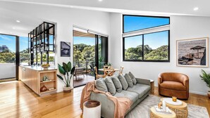 Ein modernes Wohnzimmer mit Sofa, Sessel und Couchtisch, großen Fenstern mit Blick auf die Stadt sowie einem Balkon mit Gartenmöbeln.
