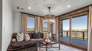 Un salón amplio con un sofá modular grande, una mesa de centro y una araña. La habitación tiene grandes puertas correderas de cristal que dan a un balcón con vistas al paisaje circundante.