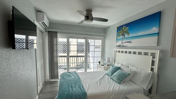 A bedroom with a bed, a ceiling fan, a painting of a beach, and a balcony with a view of the ocean. - Beach apartment, Recently Renovated, With Balcony (Clearwater)