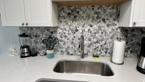 A kitchen with white cabinets, a granite backsplash, a stainless steel sink, a blender, a coffee maker, and a paper towel holder. - Beach apartment, Recently Renovated, With Balcony (Clearwater)