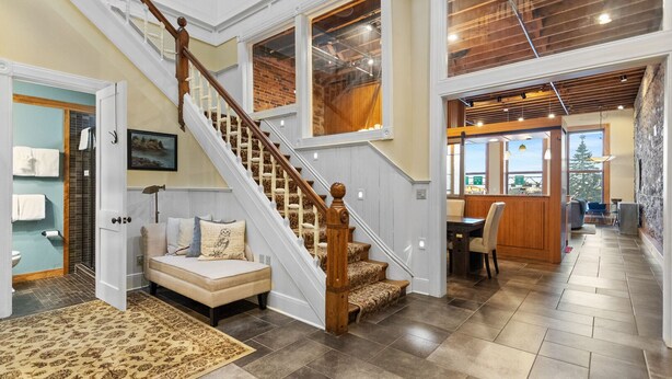 A spacious living area with a wooden staircase, a sofa with cushions, a dining table with chairs, and a view of the outdoors through large windows.