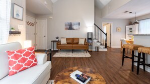 A spacious living area with a high ceiling, a brown leather sofa, a white couch with a red pillow, a round coffee table with a magazine, and a staircase leading to an upper level.