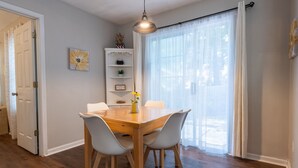 A dining area with a wooden table and four chairs, a pendant light, a white door, and a window with sheer curtains.