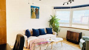 Una sala de estar moderna con un ventilador de techo, un sofá con cojines azules, una mesa de centro, una planta en maceta y vistas de la ciudad a través de amplias ventanas.