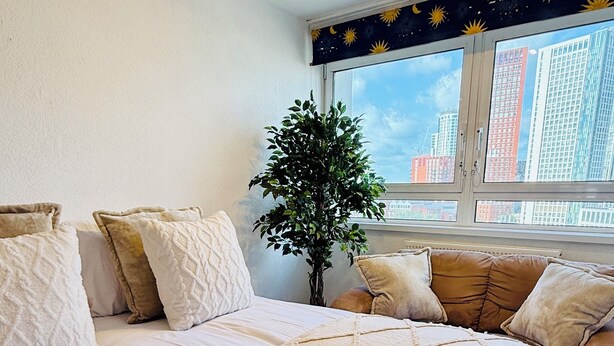 A neatly made bed with a quilted blanket and pillows, a potted plant, and a window with a view of buildings.