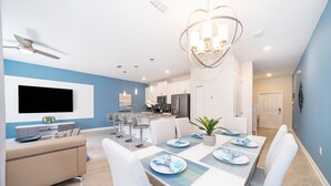 A modern dining area with a long table set for four, a TV mounted on the wall, and a ceiling fan. - LN4816 Storey Lake Home 4Beds 3Baths Private Pool and Free Waterpark (Kissimmee)