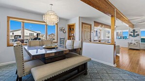 A dining area with a large table, chairs, and a bench. There are windows with a view of buildings outside. - Calypso's Dream (North) | Oceanfront | Hot Tub and Dog Friendly (Nags Head)
