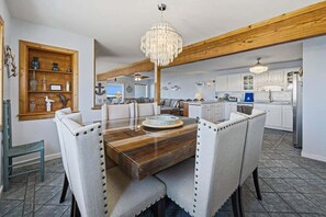 A dining area with a wooden table and upholstered chairs, a chandelier, and a kitchen in the background. - Calypso's Dream (North) | Oceanfront | Hot Tub and Dog Friendly (Nags Head)