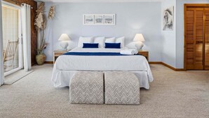 A bedroom with a bed, two bedside tables, a window with curtains, a door, and a wall with three framed pictures. - Calypso's Dream (North) | Oceanfront | Hot Tub and Dog Friendly (Nags Head)