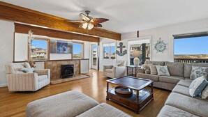 A spacious living room with a fireplace, wooden ceiling beams, and large windows offering views of the outside. - Calypso's Dream (North) | Oceanfront | Hot Tub and Dog Friendly (Nags Head)