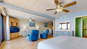 Une chambre spacieuse avec un grand lit, deux fauteuils bleus, un ventilateur de plafond et un téléviseur.