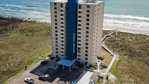Un hôtel de grande hauteur, caractérisé par un élément vertical bleu bien visible, un parking et une vue sur l’océan.