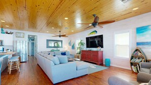 A spacious living room with a large light blue sectional sofa, wooden floors, ceiling fans, a kitchen area, and a bookshelf.