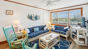Ein Wohnzimmer mit einem blau gemusterten Sofa, einem Holztisch, einem türkisfarbenen Sessel und einem Deckenventilator.